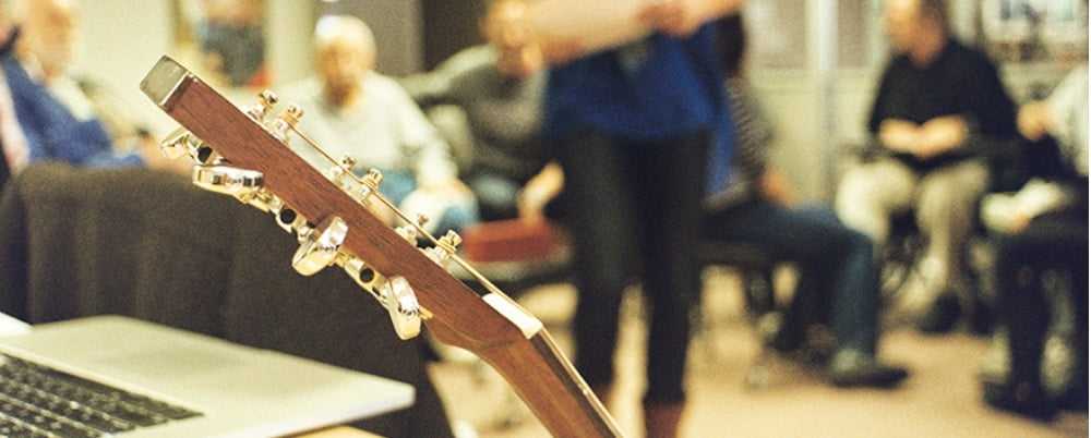 A room full of people sitting and talking with the end of a guitar in the foreground of the image